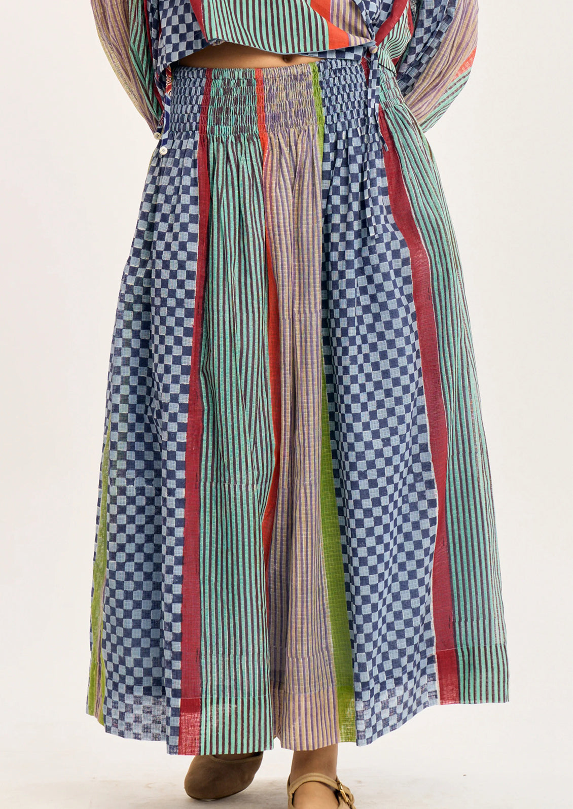 A close-up of a person wearing a long, wide-leg skirt with vertical striped and checkered fabric panels in blue, green, red, and beige tones.