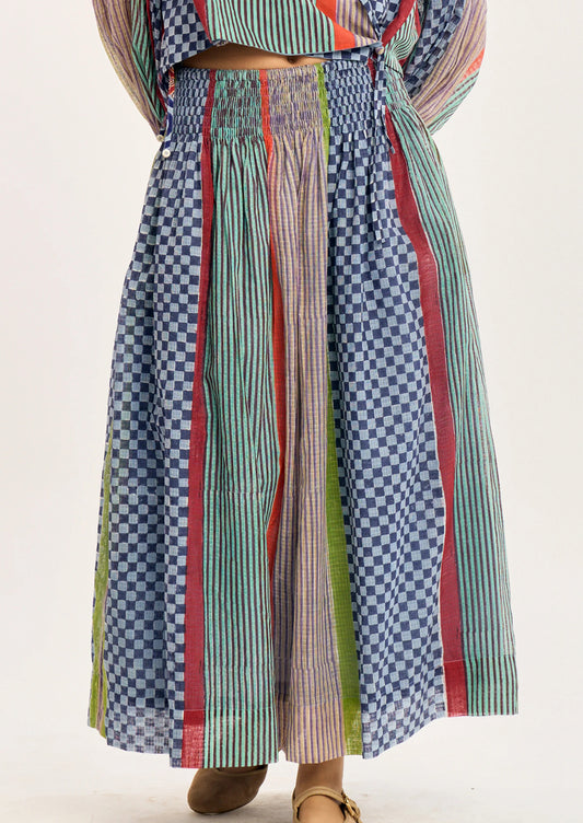A close-up of a person wearing a long, wide-leg skirt with vertical striped and checkered fabric panels in blue, green, red, and beige tones.