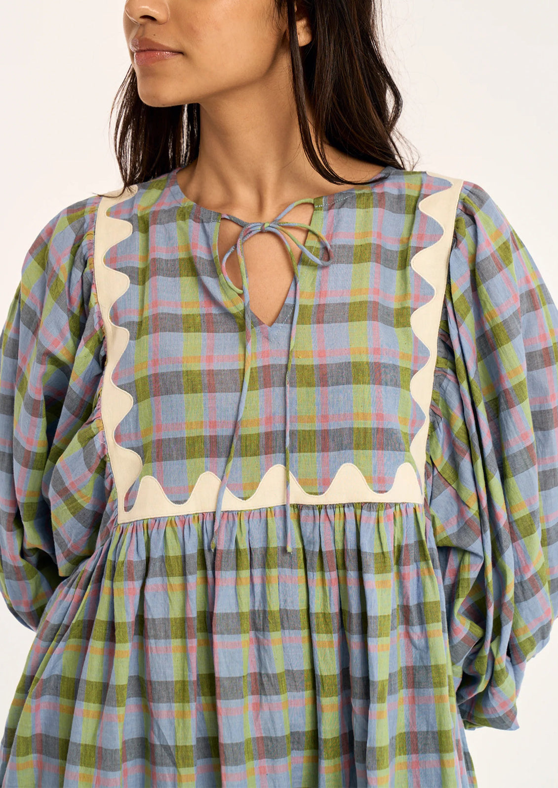 A close-up of a woman wearing a loose plaid dress with a keyhole neckline, tie detail, and scalloped cream trim over a green, blue, and pink plaid fabric.