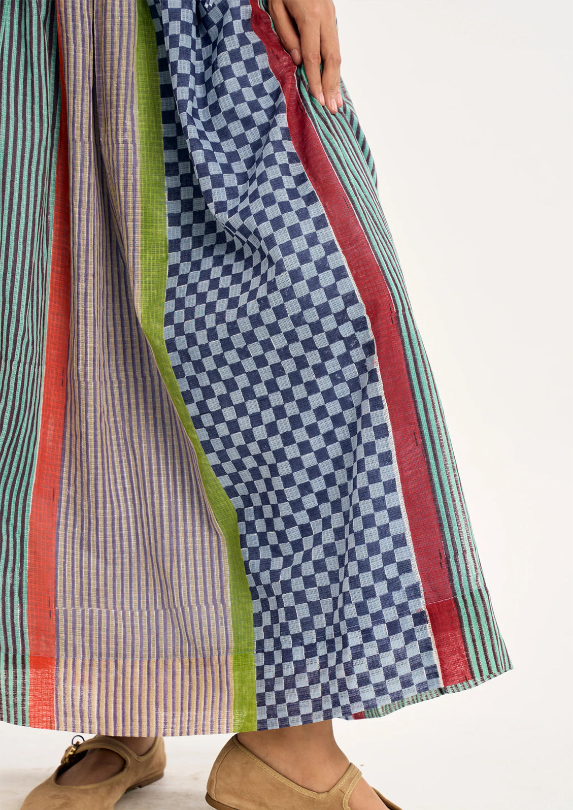 Close-up of a person wearing a long striped and checkered skirt with red, green, blue, and pink vertical borders, paired with tan flat shoes against a plain light background.