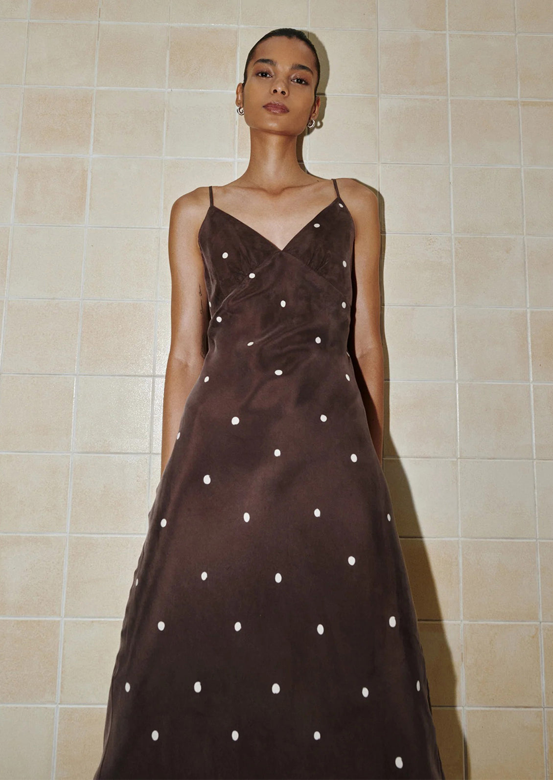 Woman wearing a brown dress with white polka dots against a beige tiled wall.