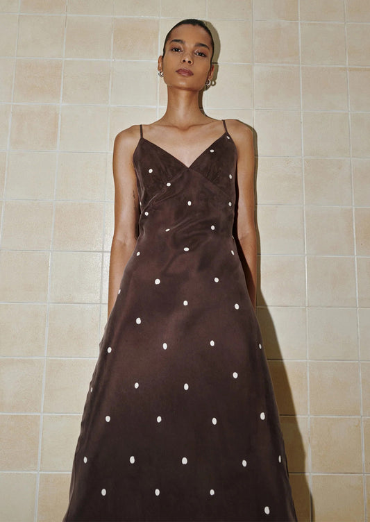 Woman wearing a brown dress with white polka dots against a beige tiled wall.