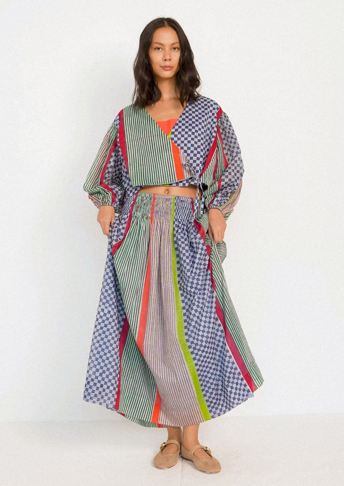 A woman modeling a matching two-piece outfit featuring a cropped long-sleeve top and a high-waisted maxi skirt with mixed patterns including stripes, checks, and solid panels in green, blue, orange, and gray.
