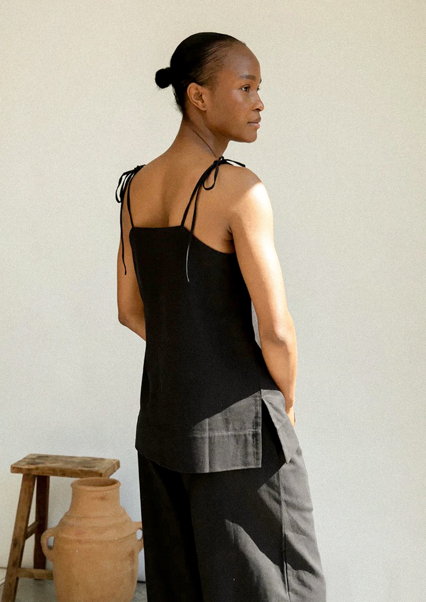 Woman wearing a black sleeveless top and gray pants against a plain wall.