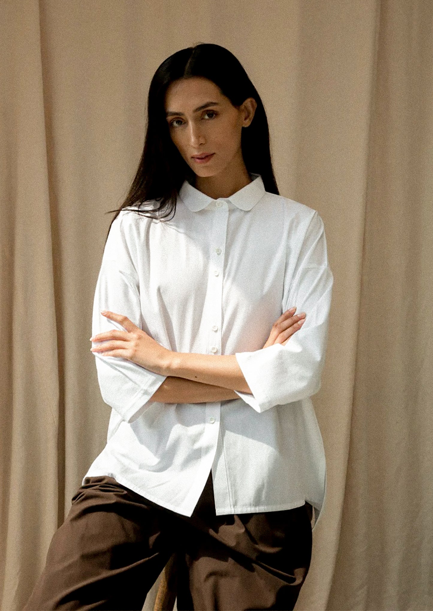 Person wearing a white shirt with arms crossed against a beige curtain background
