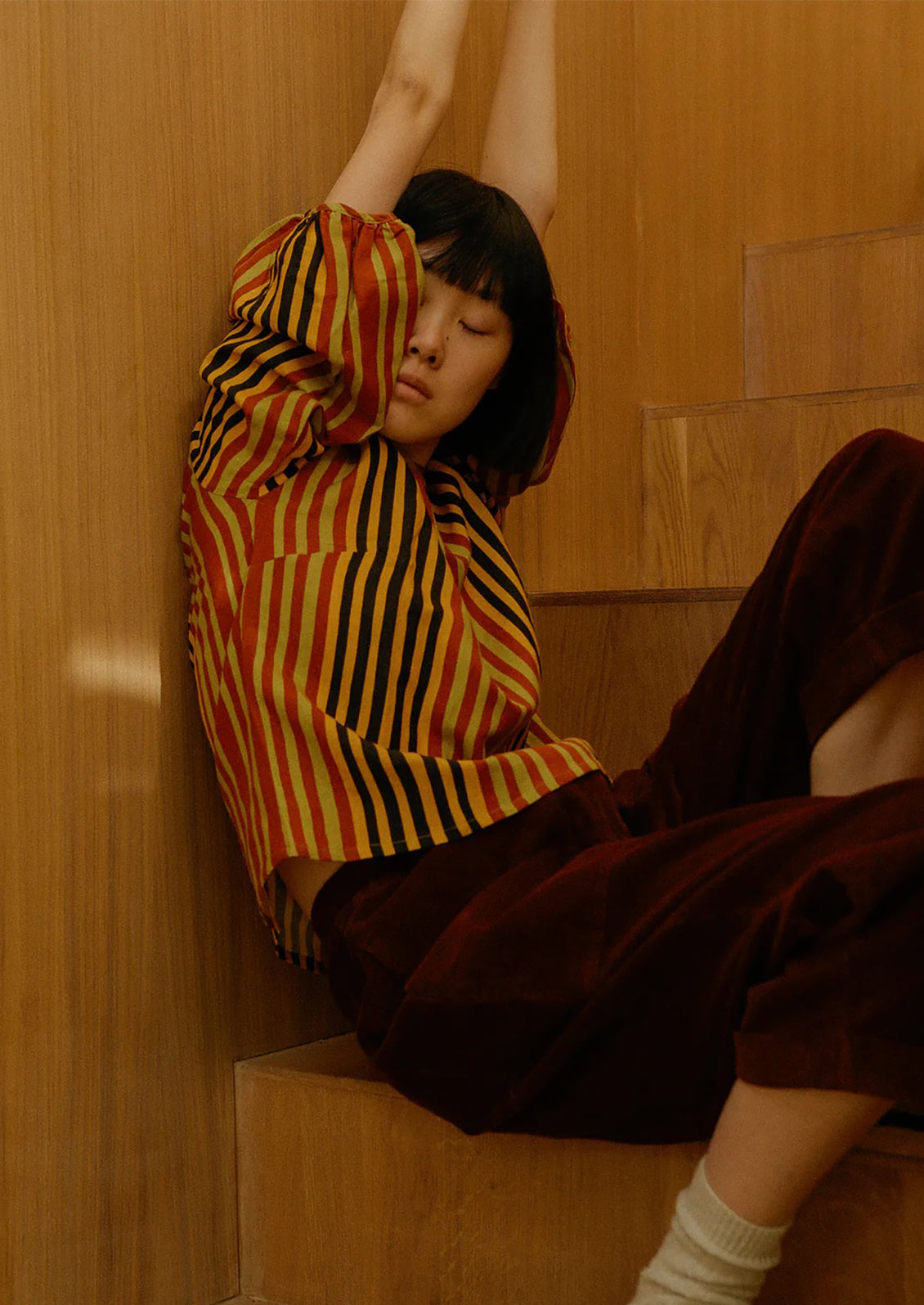 Person wearing a colorful striped shirt and brown pants sitting on a wooden staircase.
