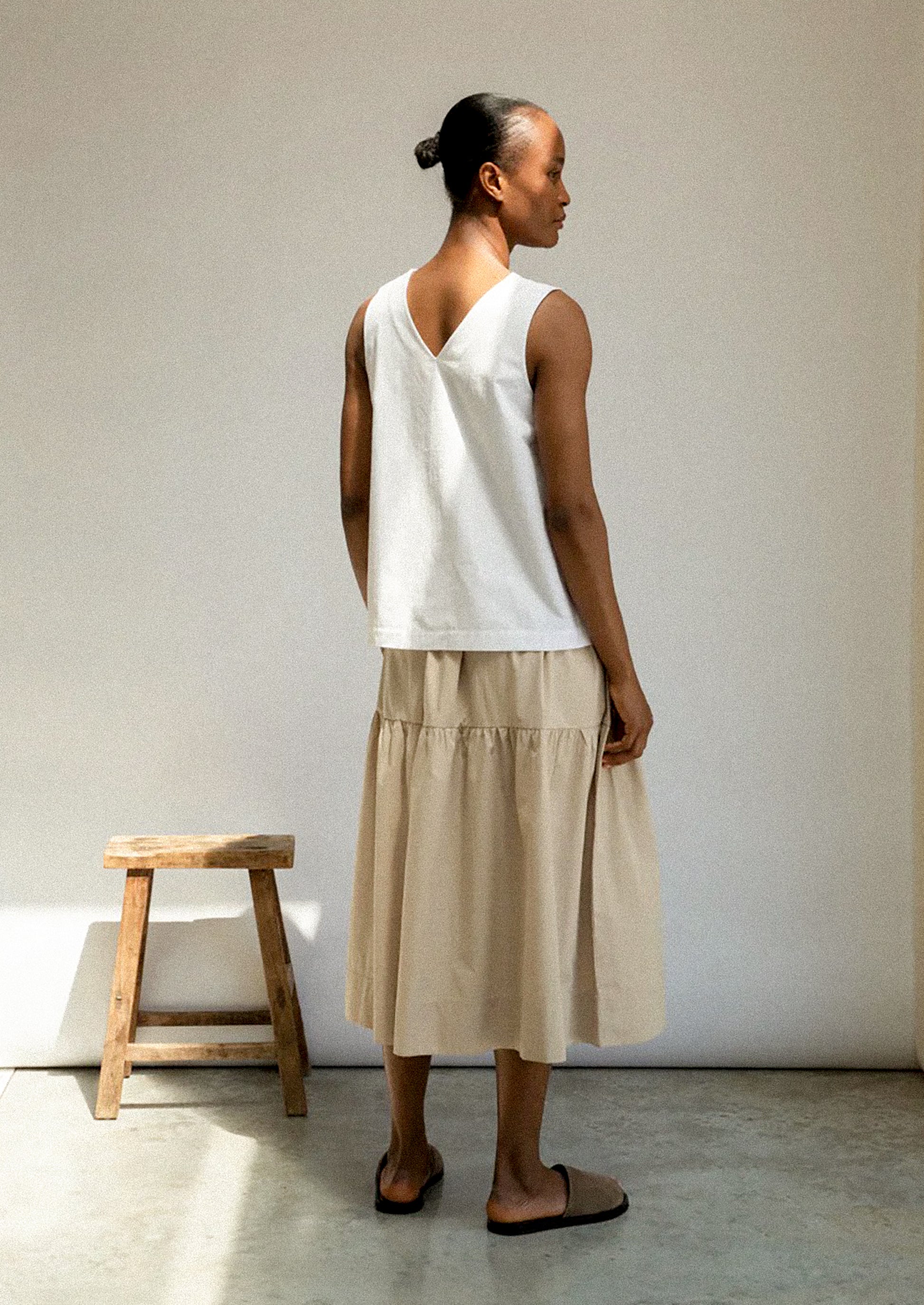 Woman wearing a white sleeveless top and beige skirt standing against a plain wall with a wooden stool.