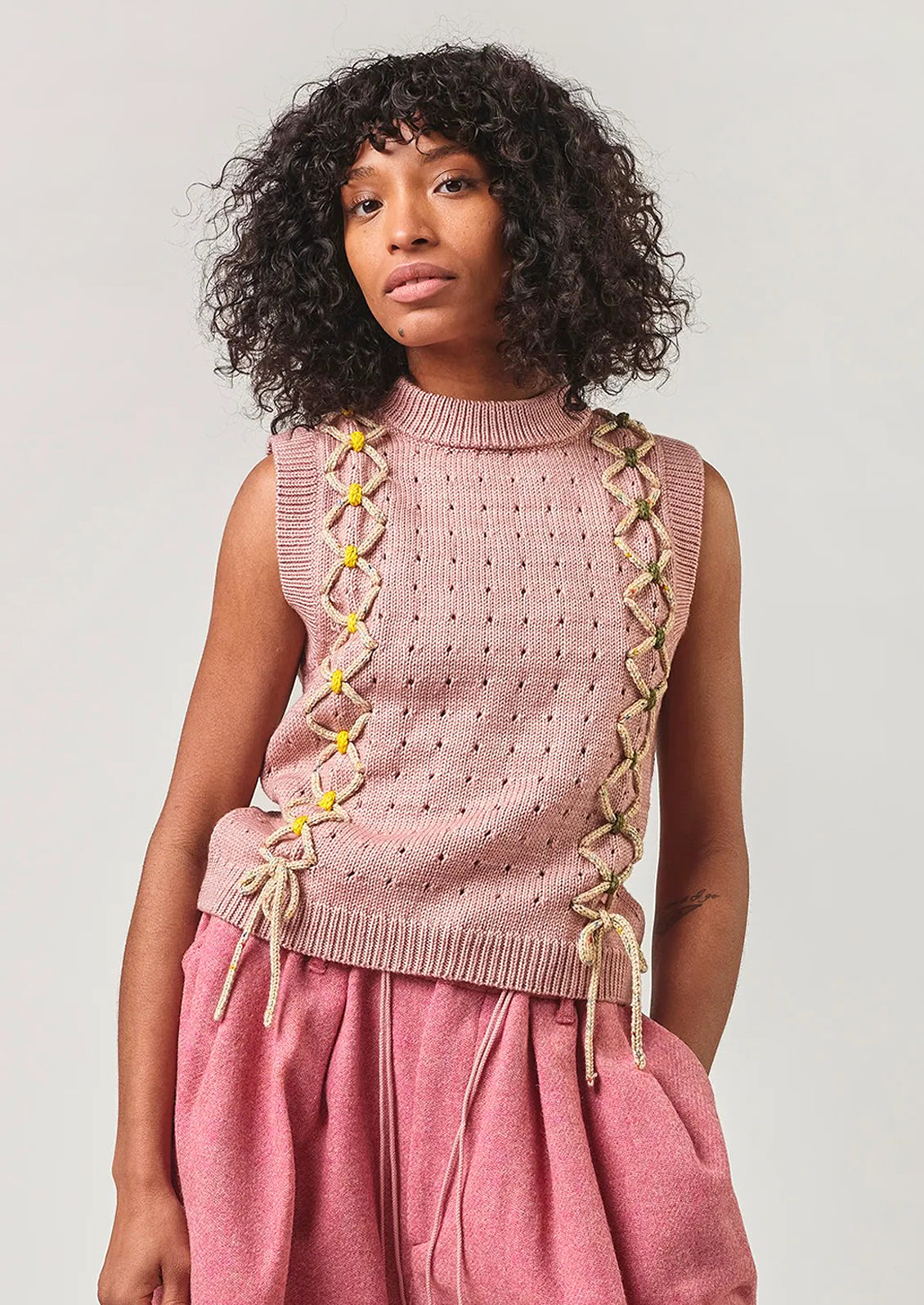 Woman wearing a pink sleeveless knitted top with decorative elements on a plain background