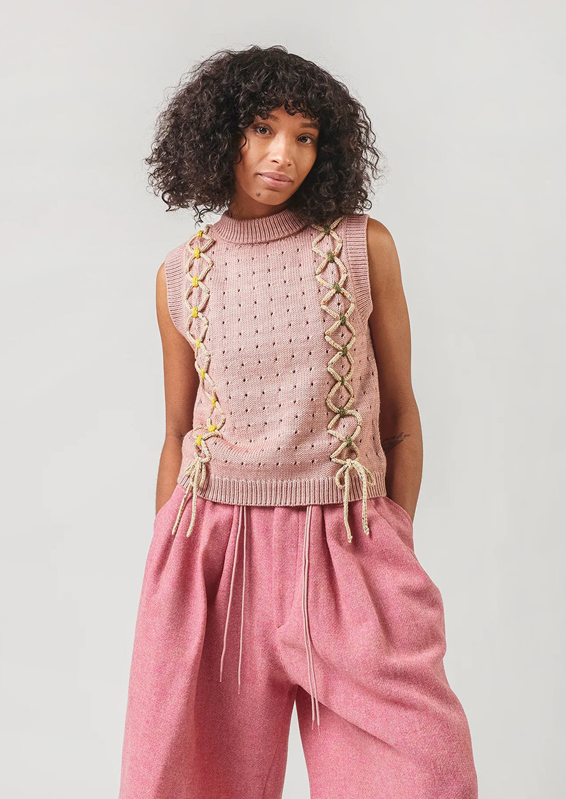 Woman wearing a pink sleeveless knitted top with lace details and pink pants against a plain background