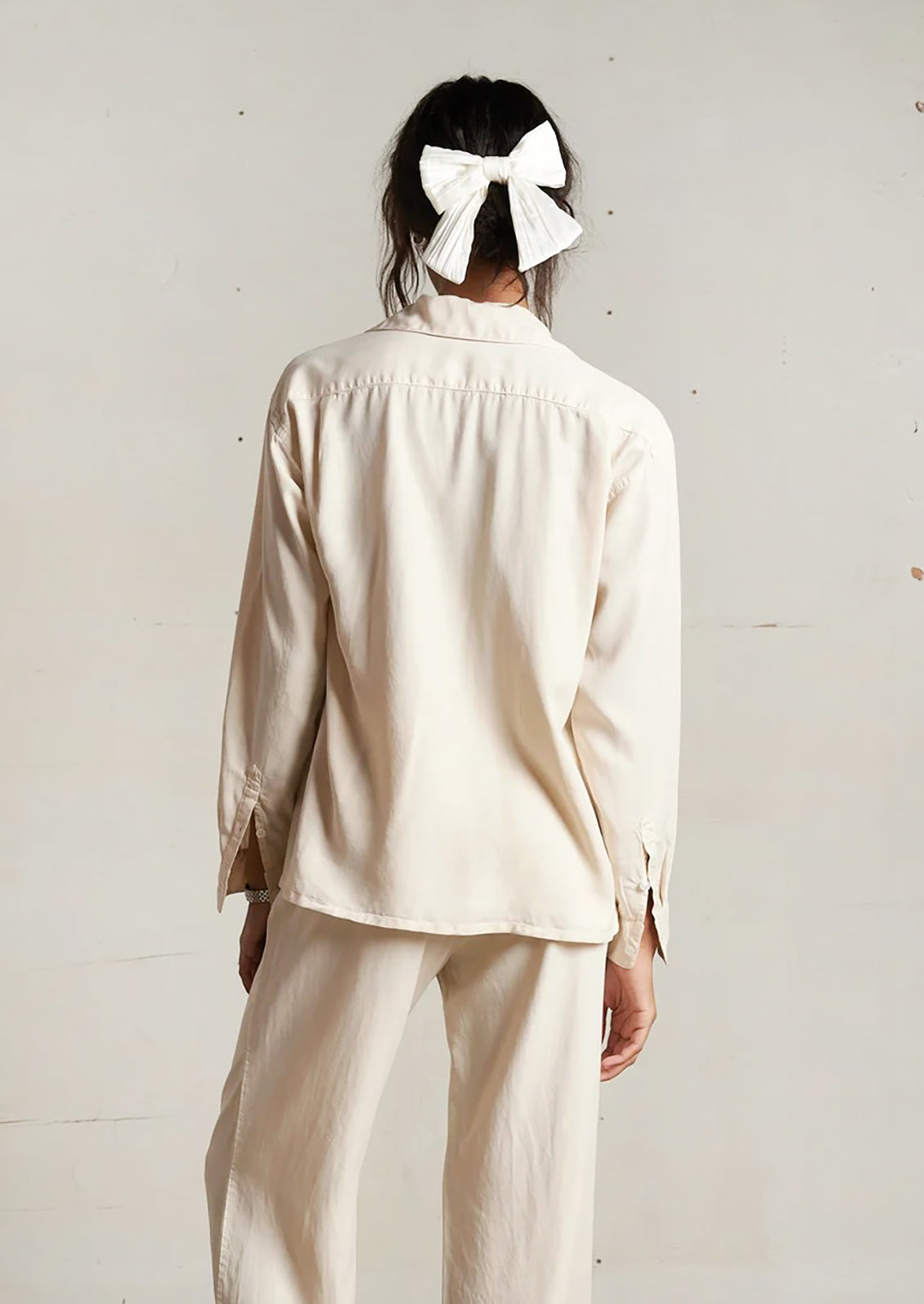 Person wearing a beige outfit with a white bow in their hair against a plain background