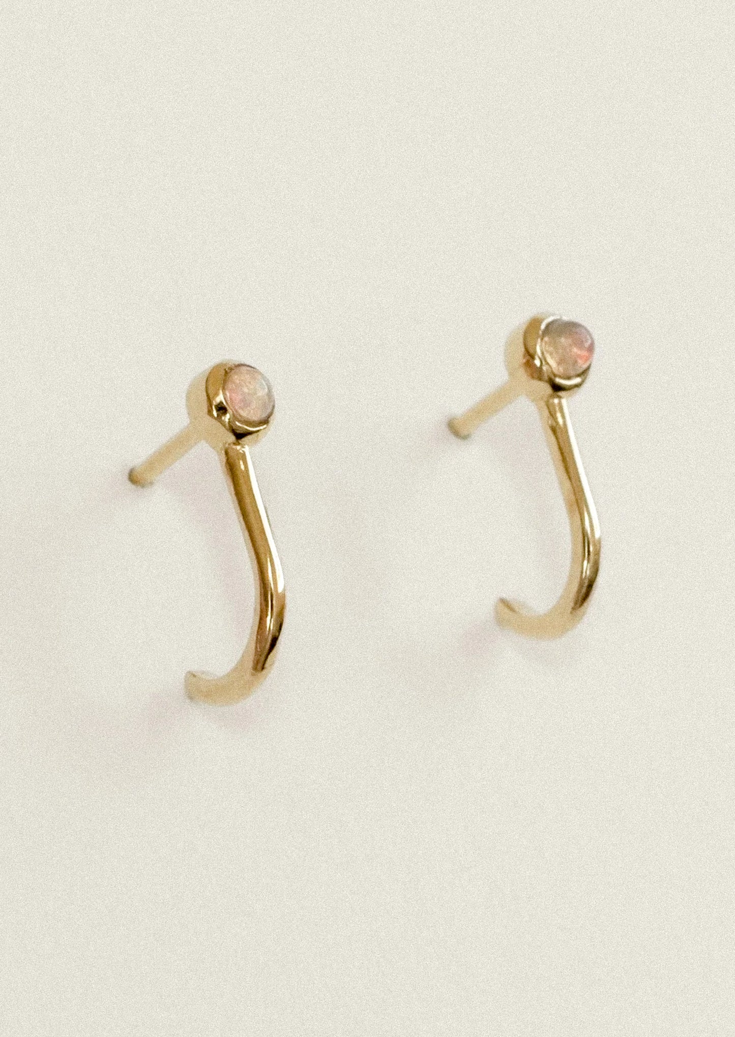 A pair of gold crescent-shaped hoop earrings with small oval pinkish-beige gemstones at the tips, displayed on a light cream background.