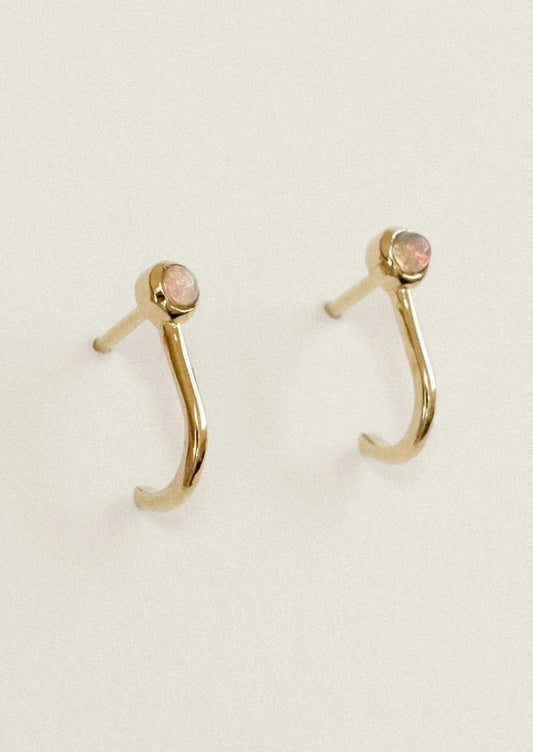 A pair of gold crescent-shaped hoop earrings with small oval pinkish-beige gemstones at the tips, displayed on a light cream background.