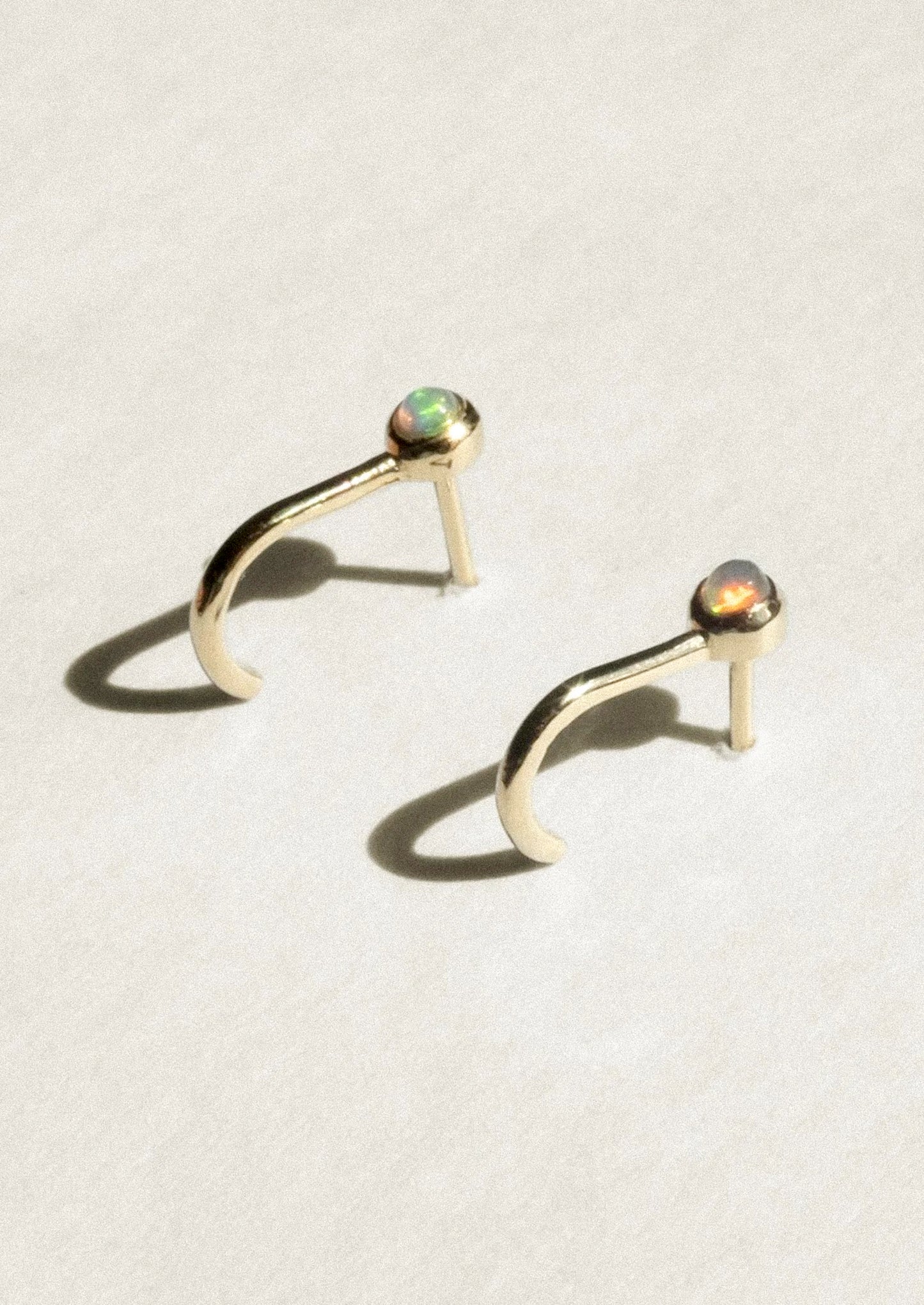A pair of gold crescent-shaped hoop earrings with small round iridescent opal gemstones at the tips, displayed on a textured off-white surface with soft shadows.