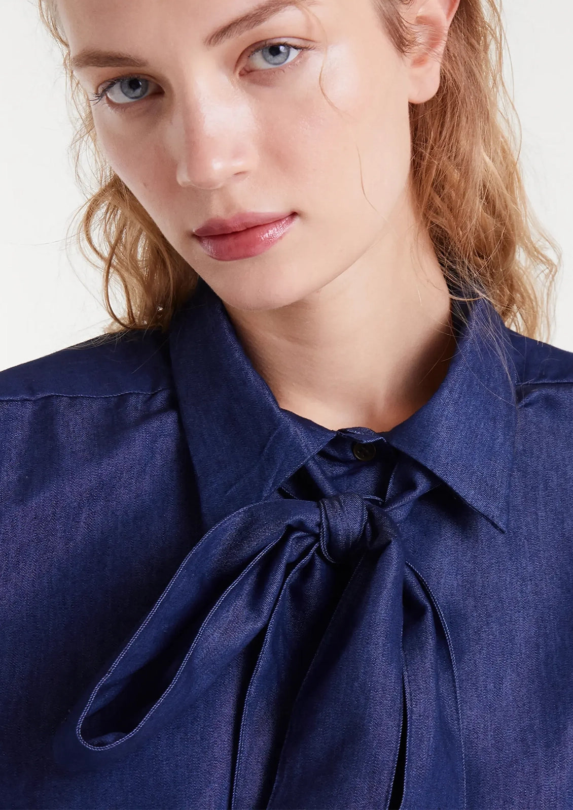 A woman wearing a dark blue chambray shirt with tie detail at neckline.