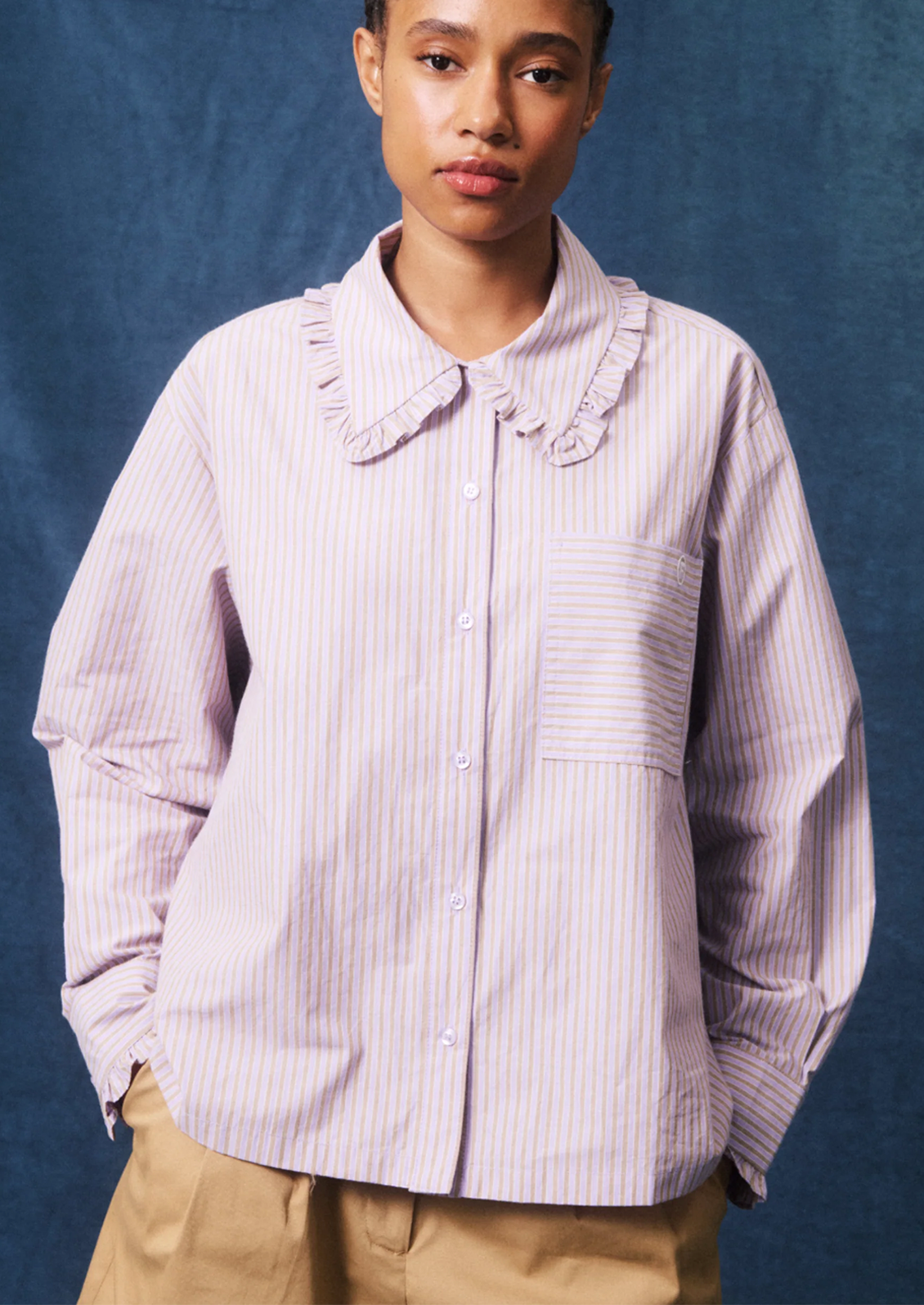 A model wearing a light pink and white vertically striped button-up shirt with a ruffled collar, paired with tan pants, set against a textured blue studio backdrop.