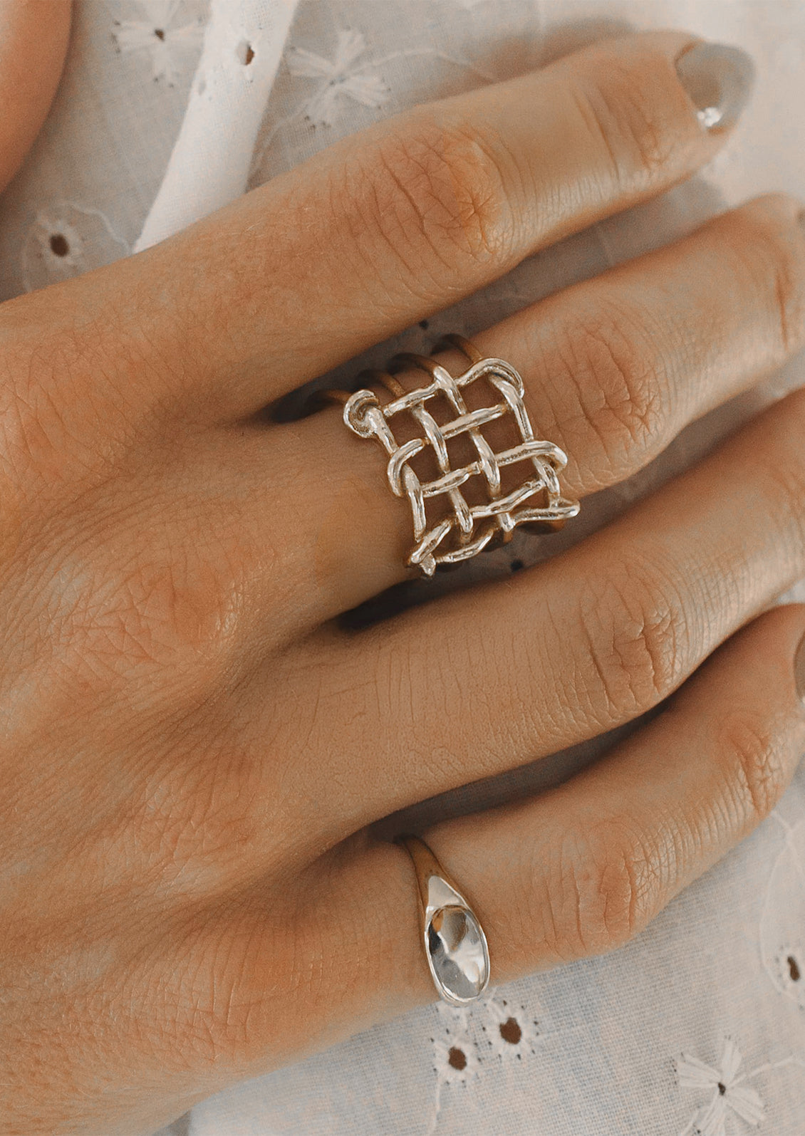 A close-up of a hand wearing a silver ring with a geometric interlaced basketweave design on the middle finger, with a smaller silver teardrop ring on the ring finger, set against a white eyelet fabric background.