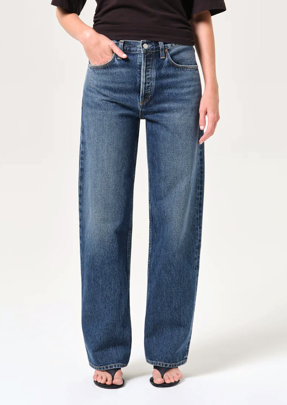 A person wearing a pair of wide-leg blue denim jeans with a black t-shirt and black sandals, shown from the waist down against a light gray studio background.