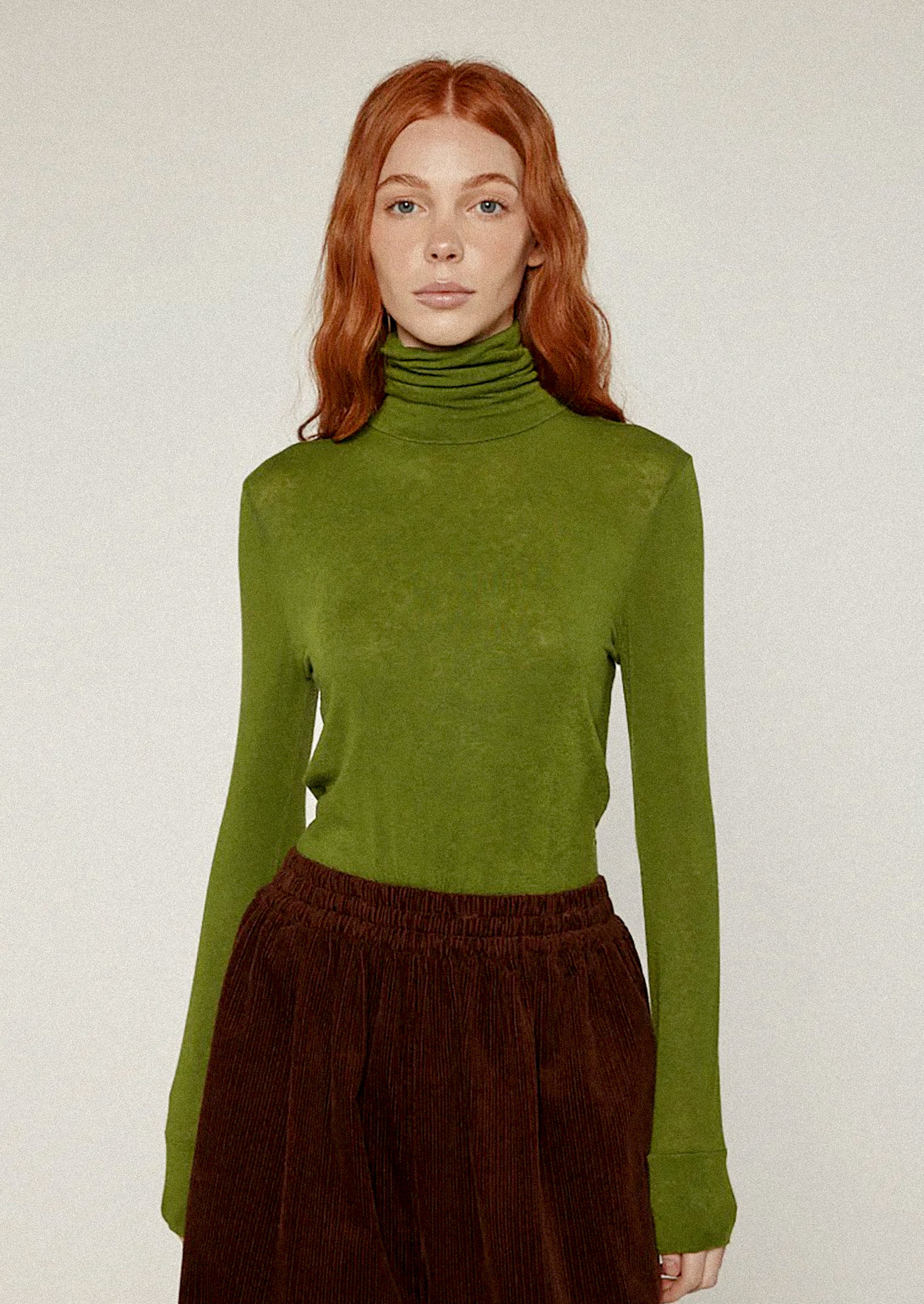 Person wearing a green turtleneck sweater and brown pants against a white background