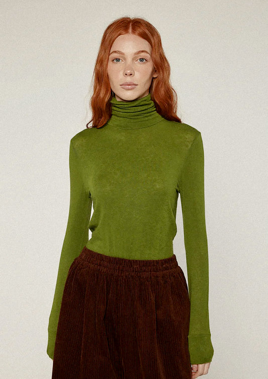 Person wearing a green turtleneck sweater and brown pants against a white background