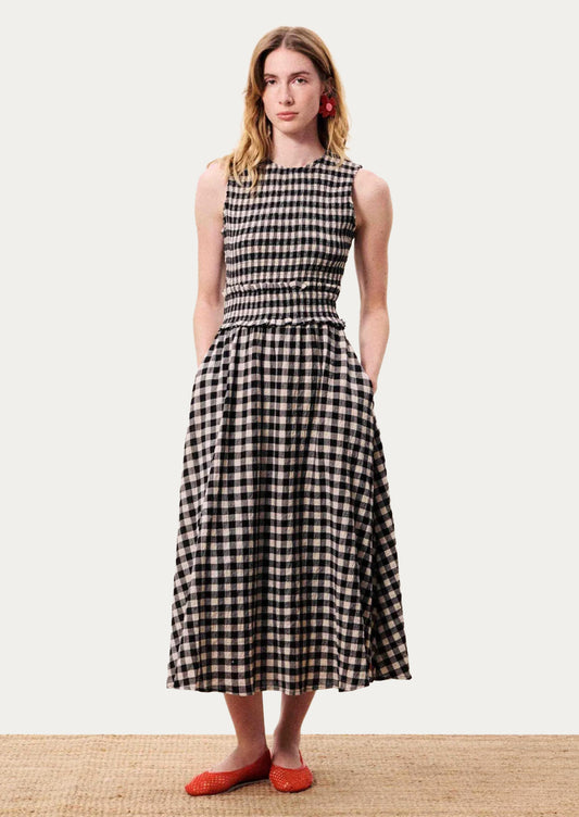 A woman modeling a sleeveless black and white gingham midi dress with a smocked waist and side pockets, paired with red shoes.