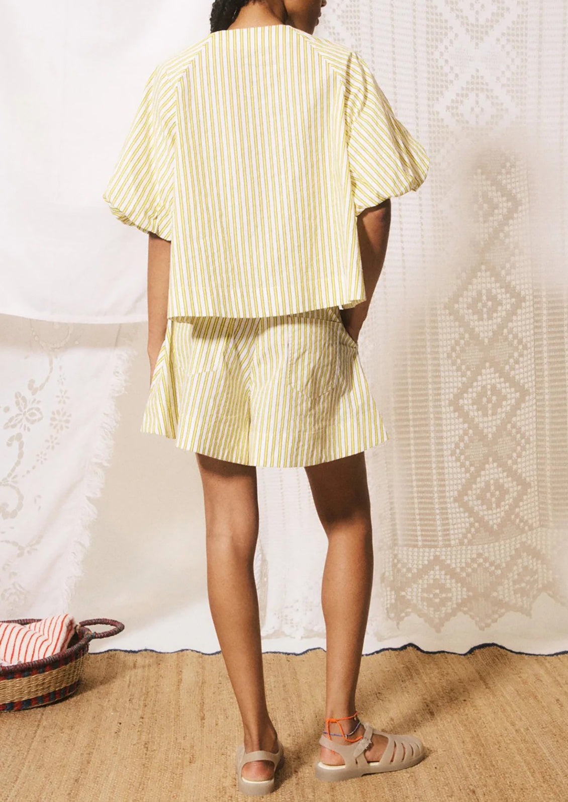 A model shown from the back wearing a light yellow and white vertically striped short-sleeve top and matching striped shorts, paired with beige sandals, standing in front of a patterned cream curtain.