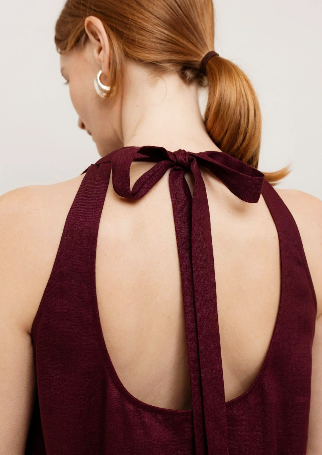 Woman wearing a burgundy dress with a halter neck and open back, standing against a plain background.
