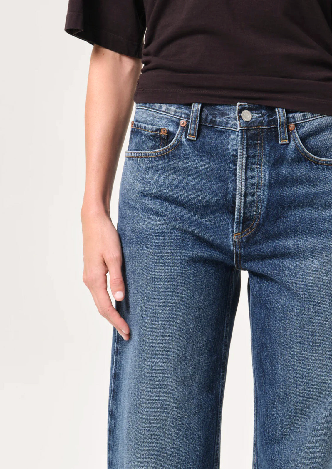 Close-up of a person wearing medium-wash blue denim jeans with a dark brown short-sleeve top, shown against a plain light background.
