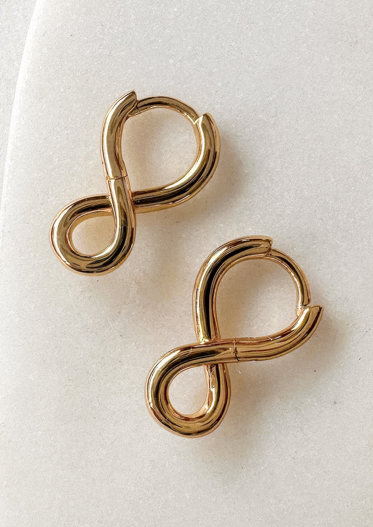 A pair of gold infinity-shaped hoop earrings on a textured off-white background.