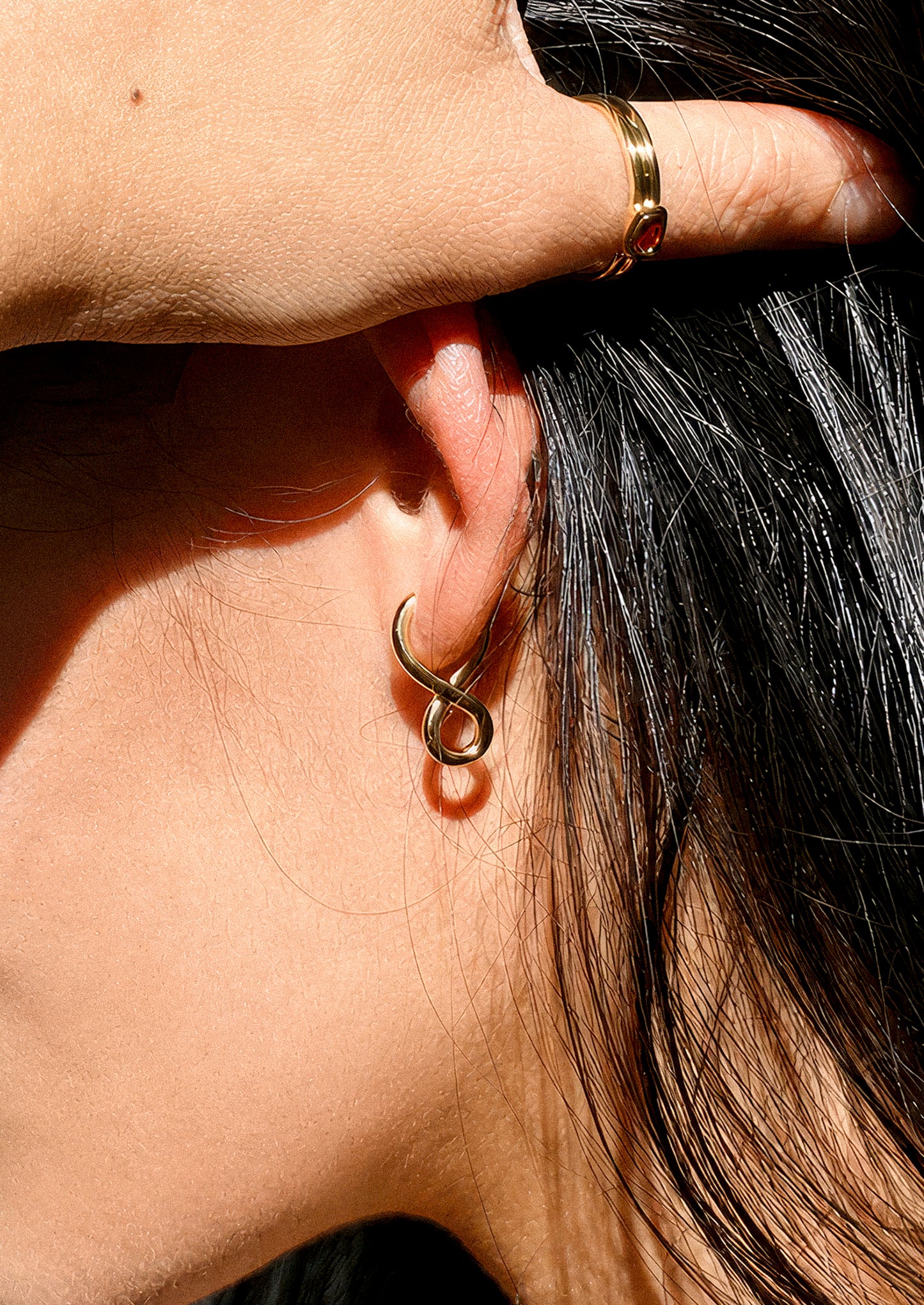 A close-up of a person's ear wearing a small infinity-shaped gold hoop earring, with a matching gold ring on the index finger. The person has dark hair and is shown in bright sunlight.