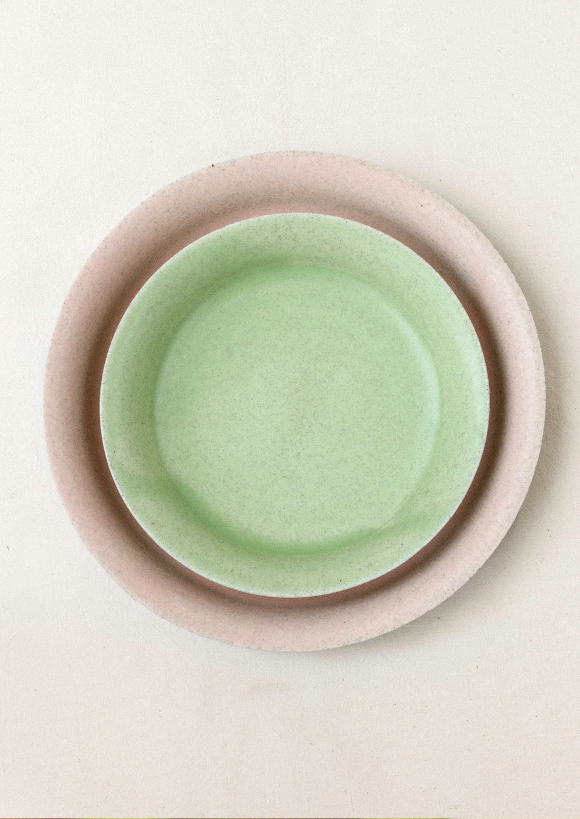 A large light pink ceramic dinner plate with a smaller green ceramic dinner plate on top set on a cream background.