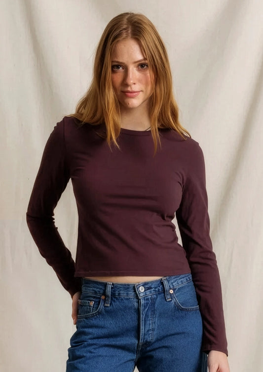 A woman modeling a fitted, long-sleeve crewneck crop top in deep plum purple, paired with blue jeans, against a draped off-white fabric backdrop.
