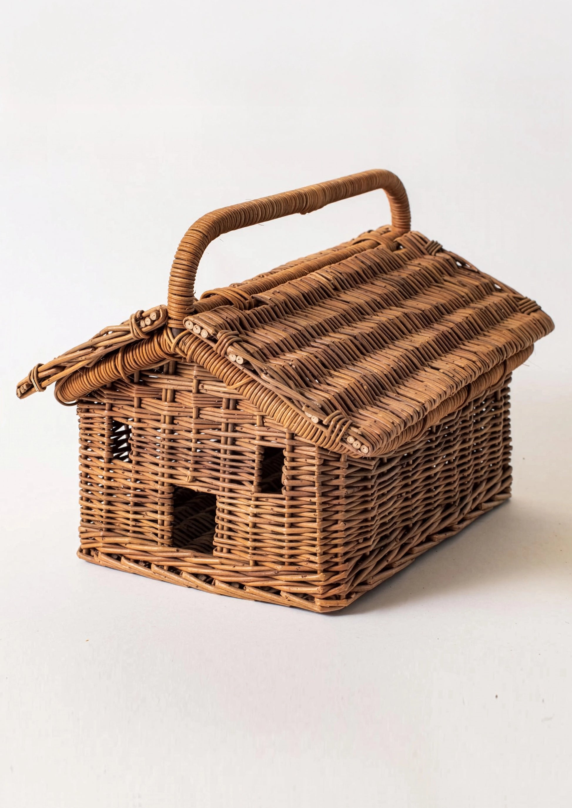 A small hand-woven wicker house-shaped basket with a hinged roof, carrying handle, and cutout windows, shown on a plain light background.
