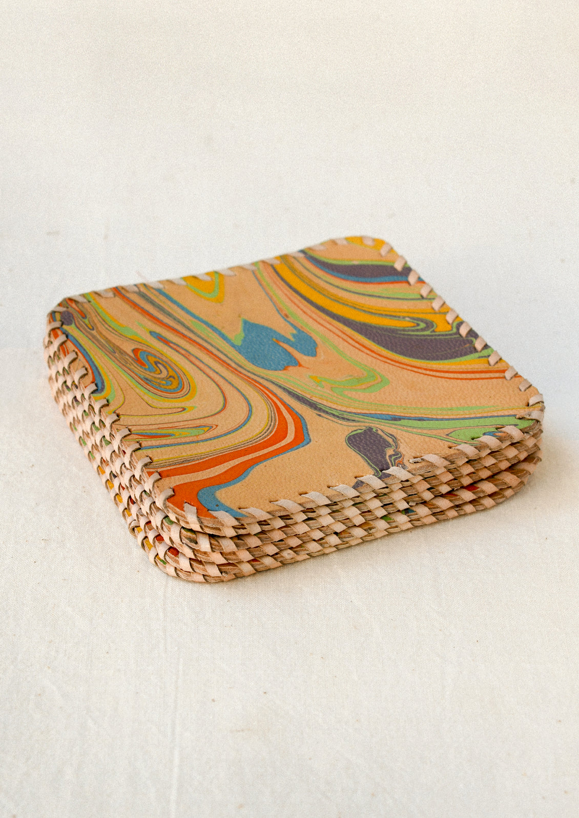 A stack of square leather coasters with rounded corners, featuring a colorful swirling marbled pattern in mustard yellow, green, blue, orange, and purple. The edges are finished with contrasting cream-colored whipstitching.