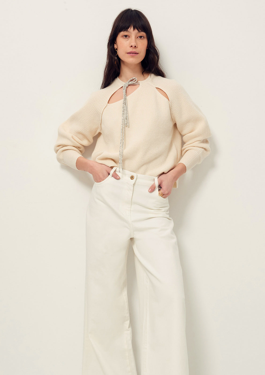 A woman wearing a cream colored sweater with cutout details and contrast tie at neck.