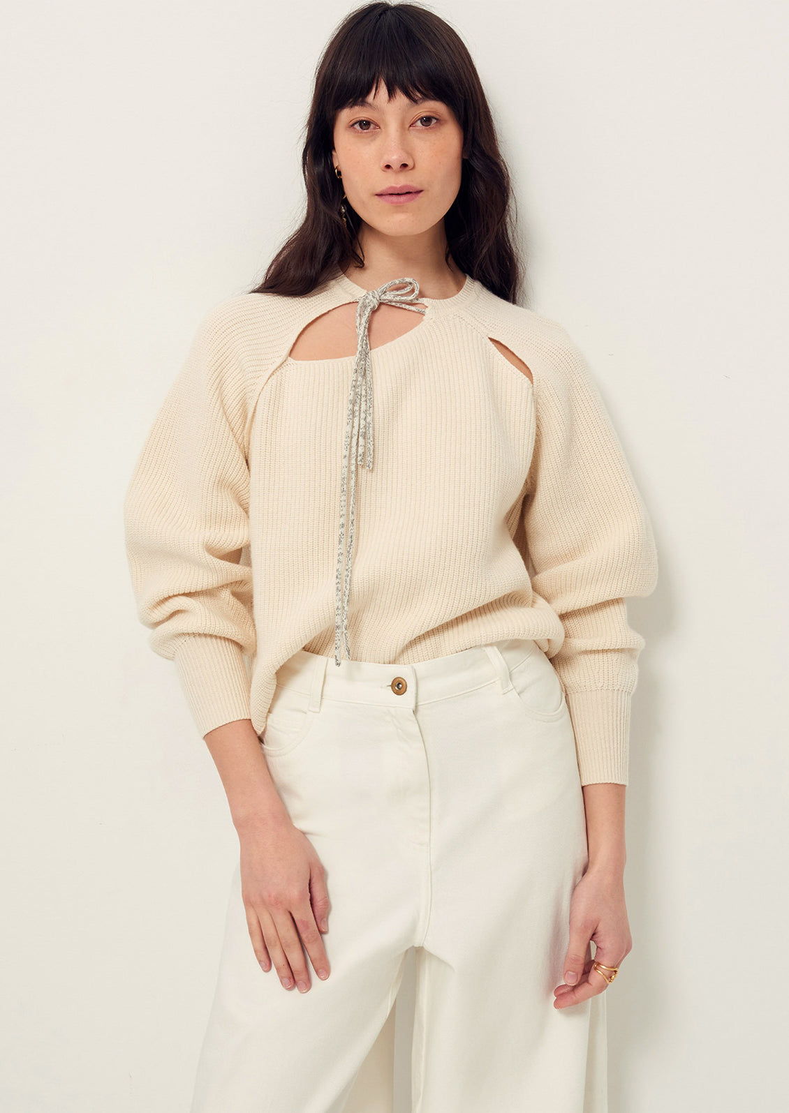 A woman wearing a cream colored sweater with cutout details and contrast tie at neck.