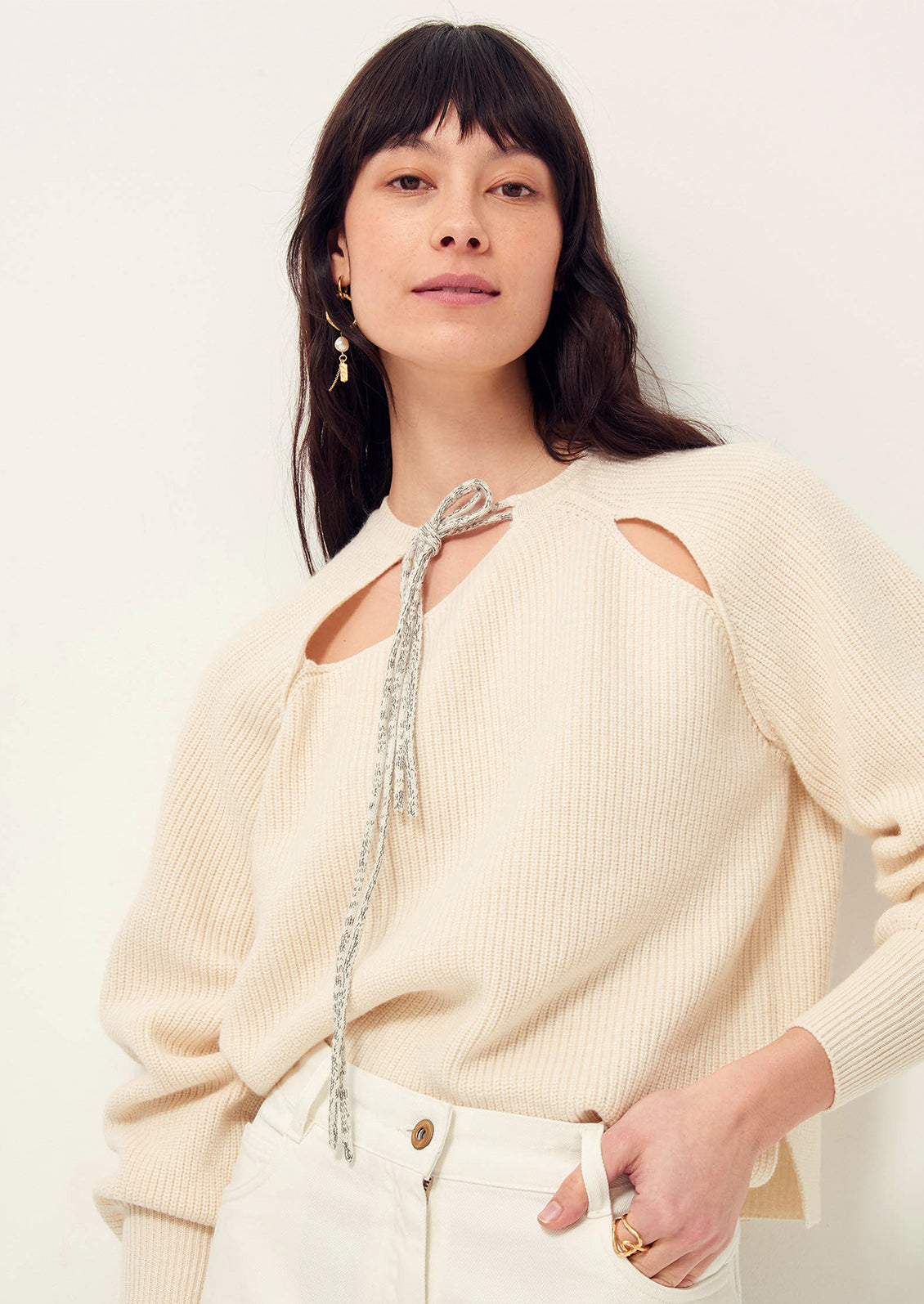 A woman wearing a cream colored sweater with cutout details and contrast tie at neck.
