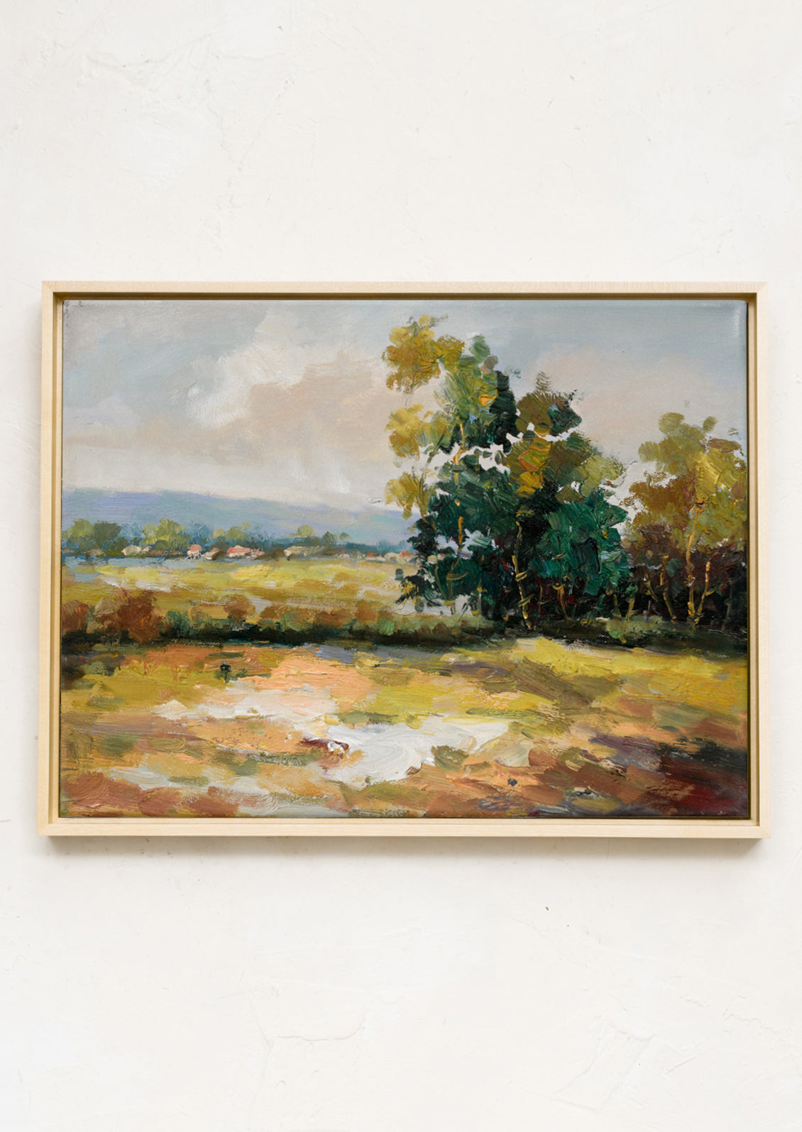 A framed original landscape oil painting of a meadow clearing with mountains and structures in the far distance.