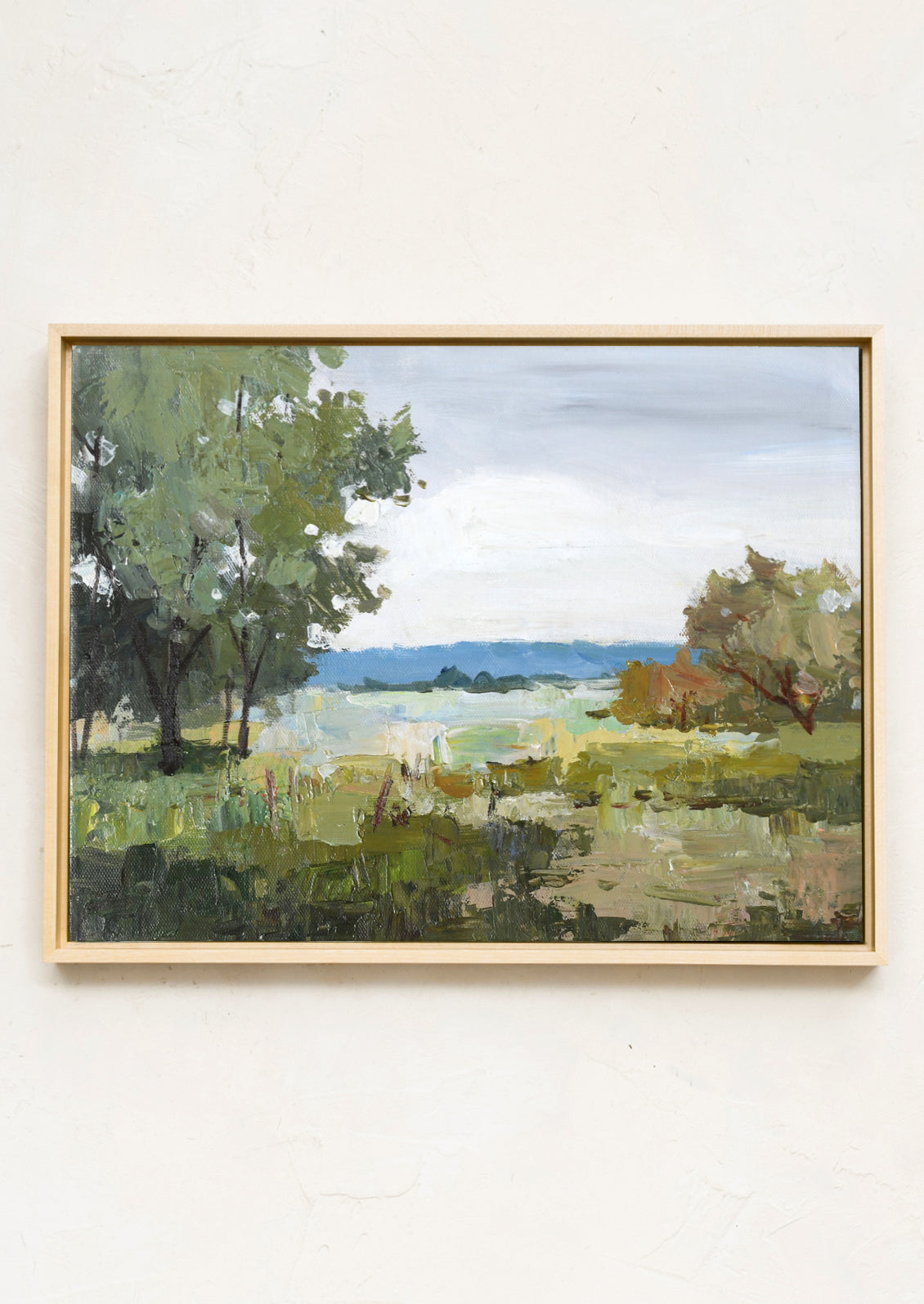 A framed landscape oil painting featuring green trees, grassy fields, and distant blue hills under a cloudy sky, housed in a light natural wood frame and mounted on a textured off-white wall.