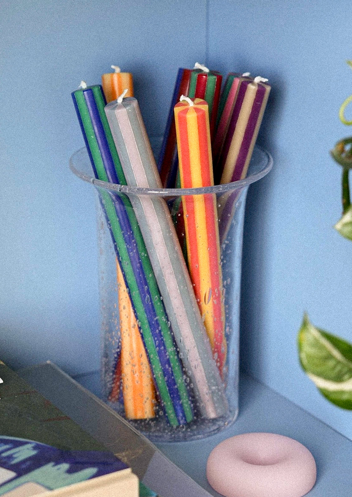 Multicolor striped taper candles in a glass vase sitting on a blue bookshelf.