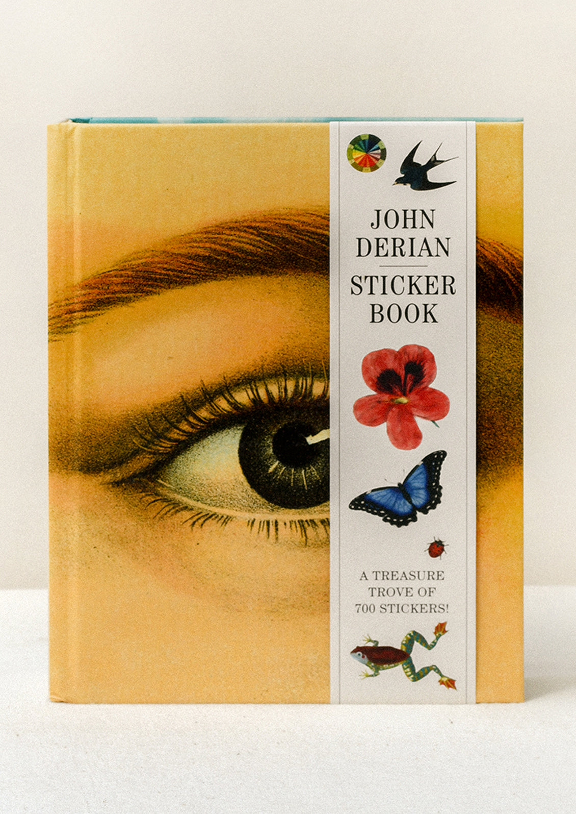 A sticker book standing on a table featuring a closeup of a woman's eye on the cover and a paper band along the side with the title "John Derian Sticker Book" and images of various animals and flowers.