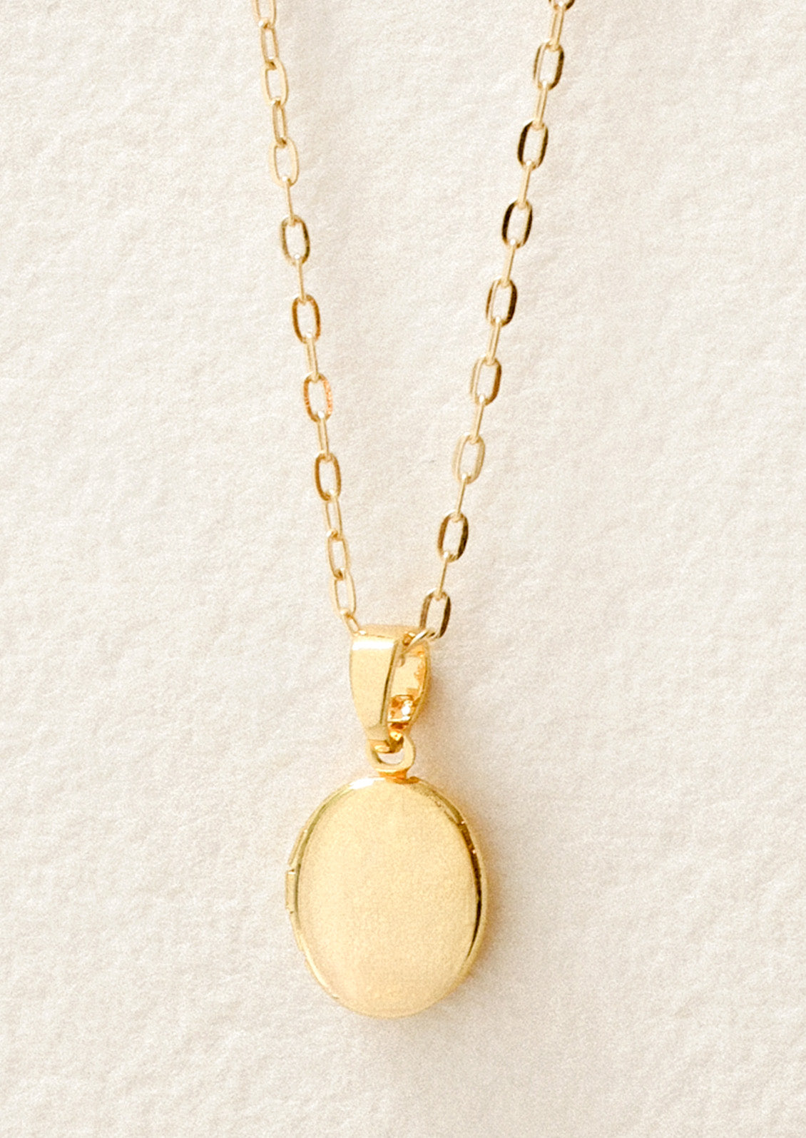 Closeup of a gold chain necklace with a small oval locket pendant on a white background.