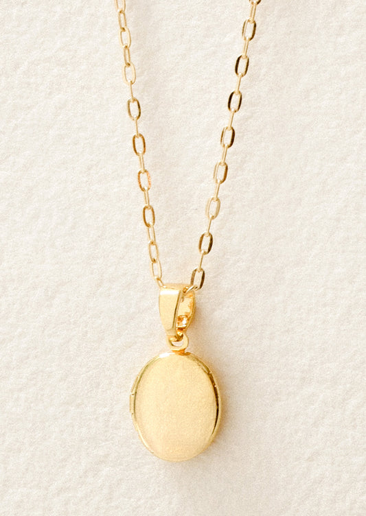 Closeup of a gold chain necklace with a small oval locket pendant on a white background.