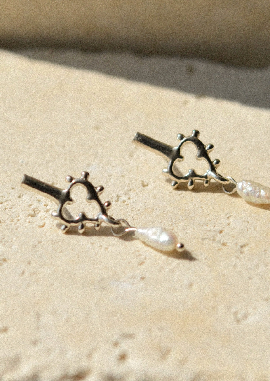 A pair of unique sterling silver earrings with a dangling keshi pearl laying on a travertine stone.
