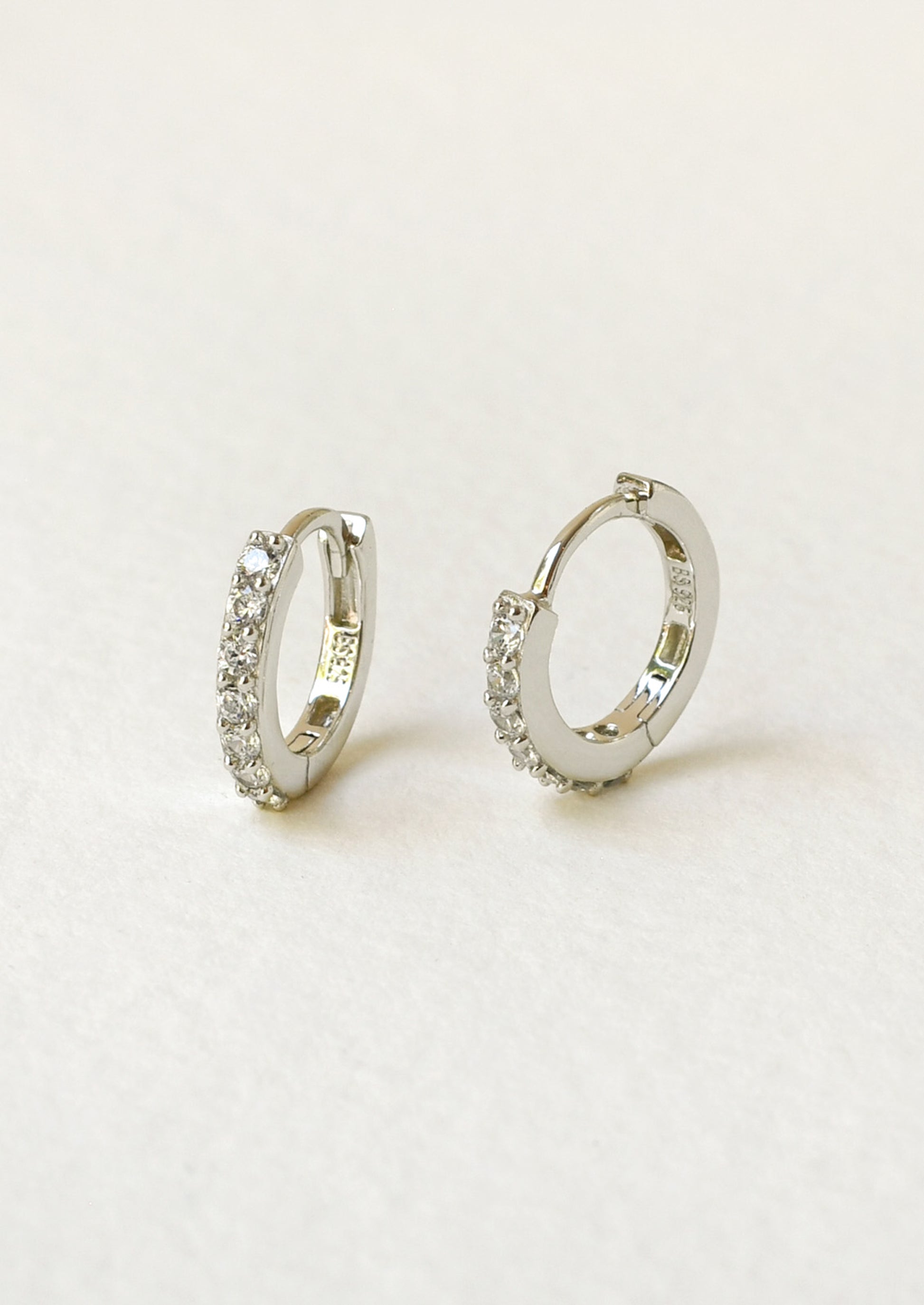 A pair of silver huggie hoop earrings adorned with small clear pavé gemstones, displayed on a clean, light beige textured surface.