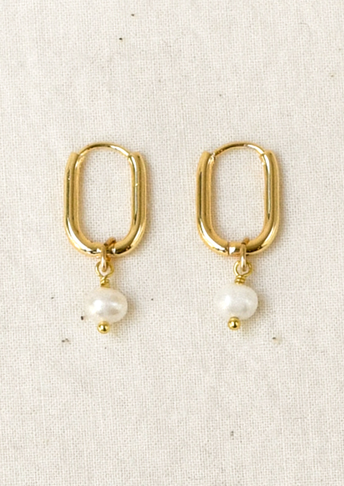 A pair of gold hoop earrings with a small white pearl dangle, displayed on a textured off-white fabric background.
