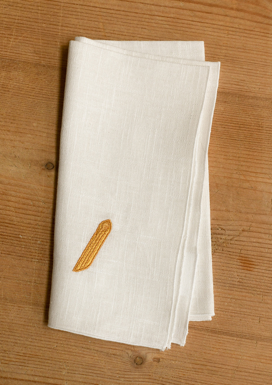 A folded white linen napkin with a small yellow embroidered noodle, displayed on a light brown wooden surface.