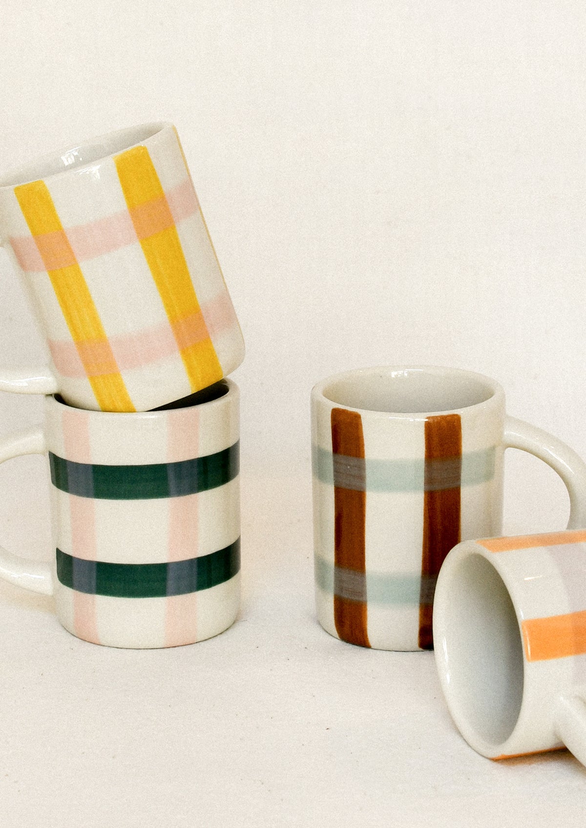 A group of ceramic mugs with colorful painted stripes and plaid patterns, including yellow, green, pink, brown, and orange on a white background.