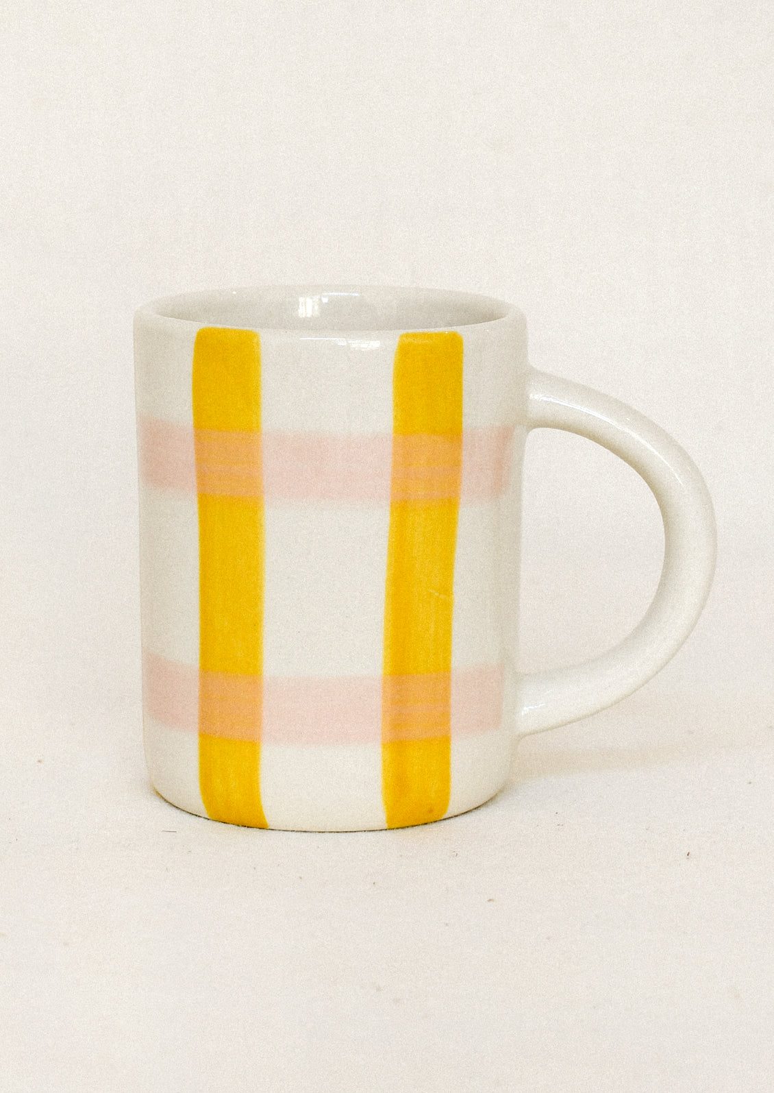 A white ceramic mug with a handle, decorated with two vertical yellow stripes and two horizontal light pink stripes, set against a plain off-white background.