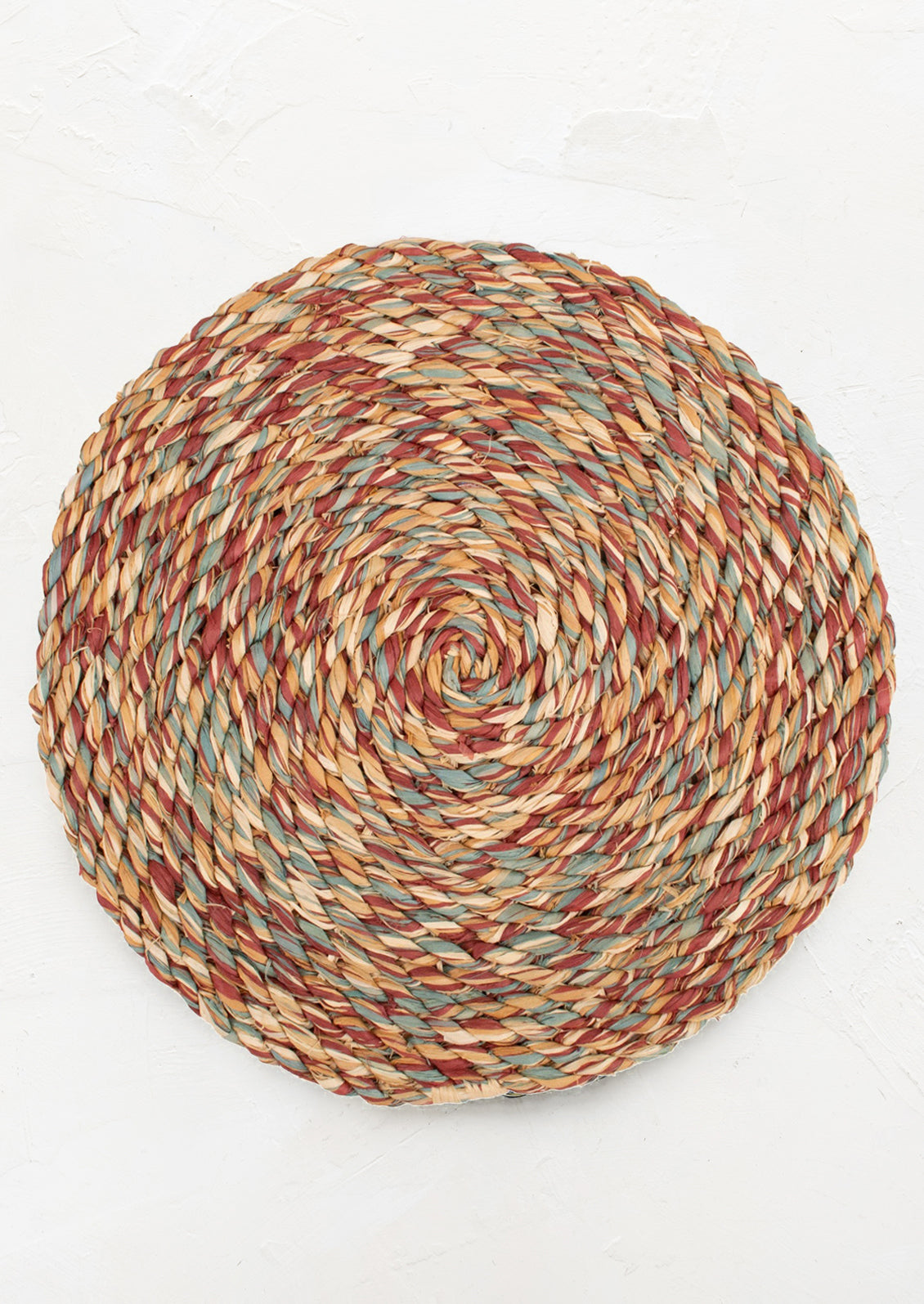 A round woven raffia placemat in multicolor rust, yellow, natural and blue.