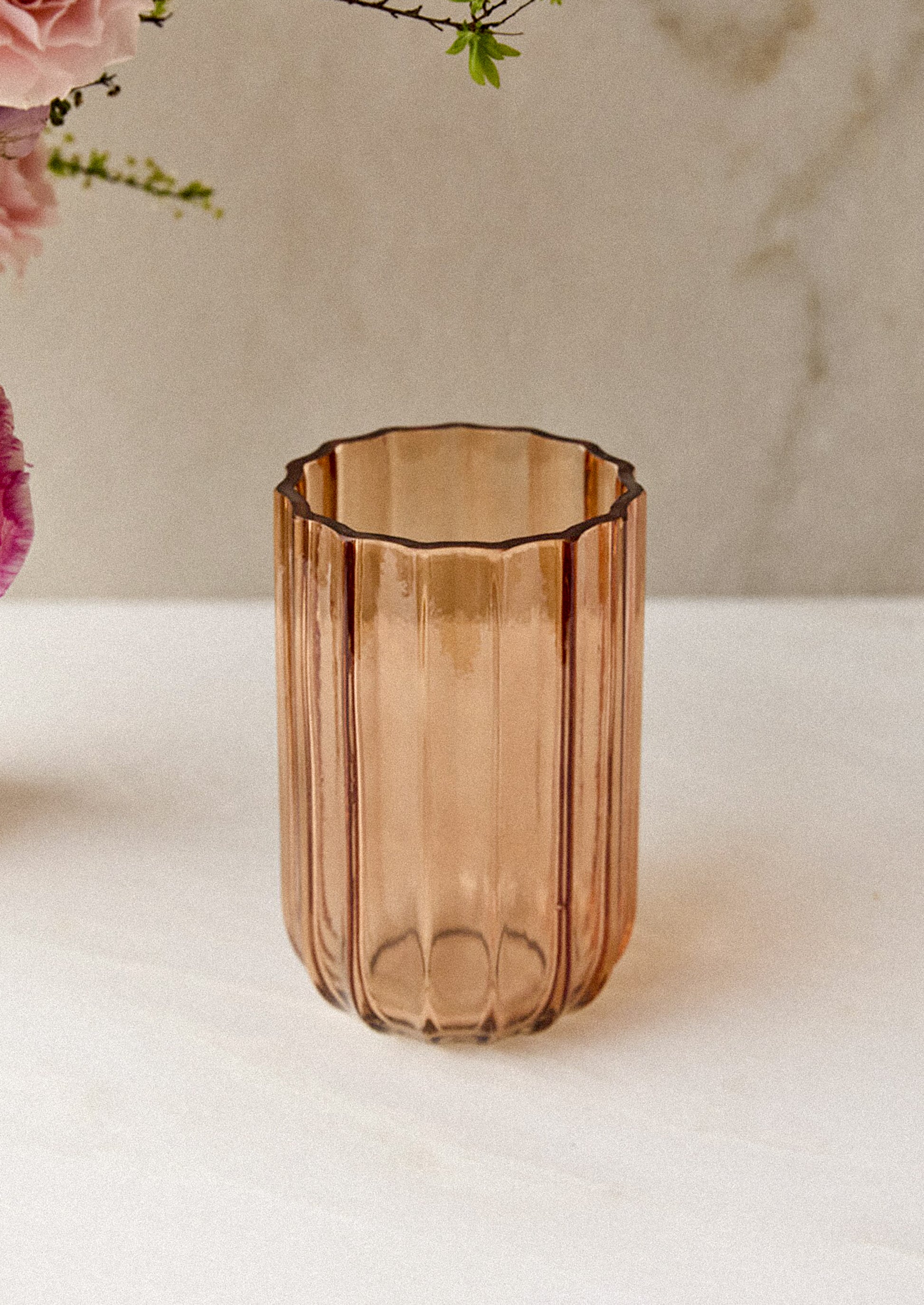 A ribbed, translucent glass vase with a scalloped rim, set on a light marble surface with soft pink flowers in the background.